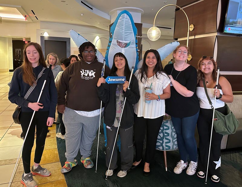A group of 2025 Summer conference attendees smiling and posing with Sharky, the JAWS mascot.